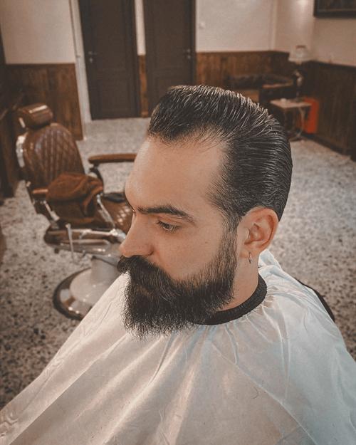 Άντρας με μαλλιά χτενισμένα προς τα πίσω και πυκνή σκούρα γενειάδα, με το μωσαϊκό και μια παλιά πολυθρόνα κουρείου πίσω του